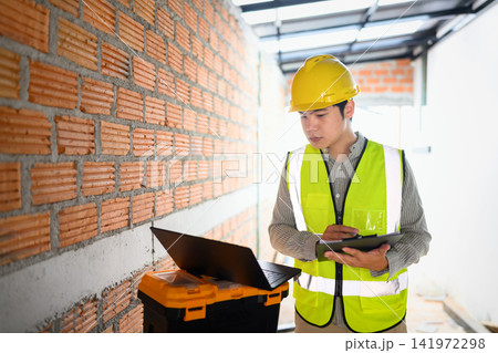 Young Asian Engineer in Yellow Hard Hat and Safety Vest Reviewing Clipboard at Construction Site with Brick Wall Young Asian Engineer in Yellow Hard Hat and Safety Vest Reviewing Clipboard at Construction Site with Brick Wall 141972298