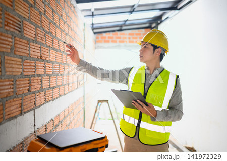 Asian Construction Engineer Inspecting Brick Wall at Building Site Wearing Safety Vest and Hard Hat with Clipboard 141972329
