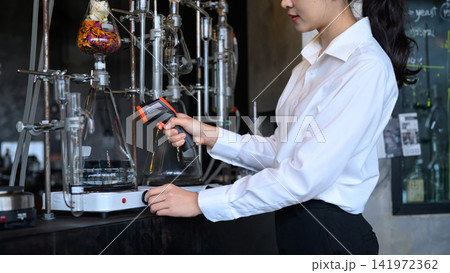 Young Asian Woman Using Infrared Thermometer on Laboratory Glassware During Botanical Distillation Process Young Asian Woman Using Infrared Thermometer on Laboratory Glassware During Botanical Distillation Process 141972362