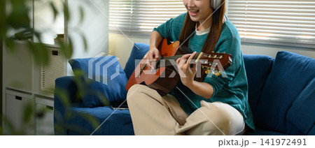 Closeup Woman Playing Acoustic Guitar with Headphones on Blue Sofa at Home 141972491