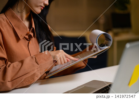 Young Asian Woman Reading Magazine at Desk with Laptop in Modern Office Young Asian Woman Reading Magazine at Desk with Laptop in Modern Office 141972547