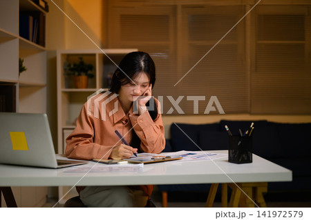 Young Asian Gen Z Business Woman Working Late at Home Office Reviewing Documents with Laptop 141972579
