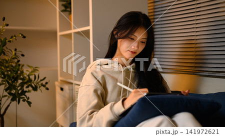 Young Asian Woman Using Digital Tablet with Stylus Pen at Home in Cozy Living Room Evening Light 141972621