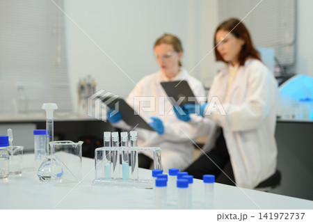 Two Female Scientists in Lab Coats Reviewing Data with Test Tubes and Beakers in Foreground 141972737