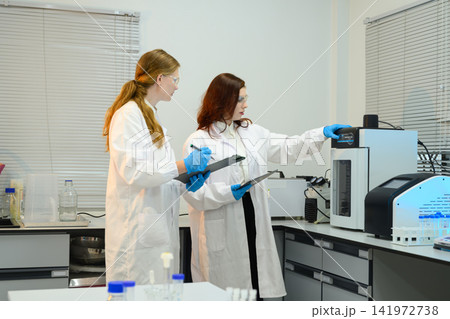Two Female Scientists in Lab Coats Operating Laboratory Equipment While Recording Data on Clipboard Two Female Scientists in Lab Coats Operating Laboratory Equipment While Recording Data on Clipboard 141972738