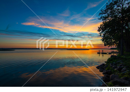 Colorful Sunset over Calm Lake with Wooden Pier and Two People Standing on the Dock Colorful Sunset over Calm Lake with Wooden Pier and Two People Standing on the Dock 141972947
