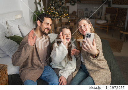 Merry Christmas celebrating. Happy family mother father girl child having video chat in smartphone. Parents kid recording video in social network. Family having video call with friends family Merry Christmas celebrating. Happy family mother father girl child having video chat in smartphone. Parents kid recording video in social network. Family having video call with friends family 141972955