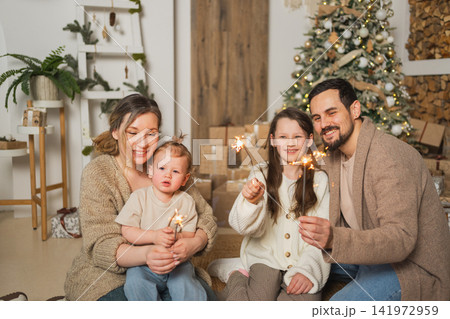 Merry Christmas Happy family mother father two children with glittering sparklers candle burns near Christmas tree. Parents kids celebrating Christmas enjoying winter holidays together. Family moments Merry Christmas Happy family mother father two children with glittering sparklers candle burns near Christmas tree. Parents kids celebrating Christmas enjoying winter holidays together. Family moments 141972959