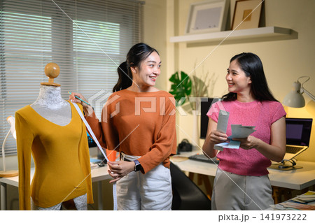 Two Asian Female Fashion Designers Collaborating in Studio, Measuring Clothing on Mannequin Together Two Asian Female Fashion Designers Collaborating in Studio, Measuring Clothing on Mannequin Together 141973222