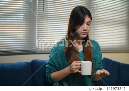 Young Asian Woman Hesitating to Take Medicine Pills While Holding a Mug at Home Young Asian Woman Hesitating to Take Medicine Pills While Holding a Mug at Home 141973241