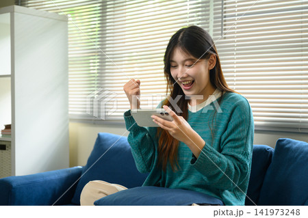 Excited Young Asian Woman Celebrating Good News on Smartphone While Sitting on Sofa at Home Excited Young Asian Woman Celebrating Good News on Smartphone While Sitting on Sofa at Home 141973248