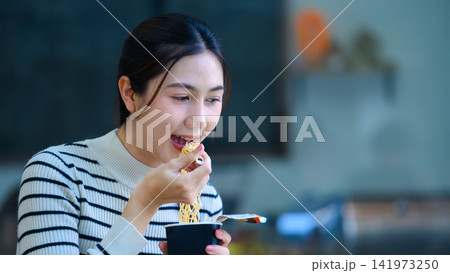 Young Asian Woman Eating Instant Noodles from Cup with Focused Enjoyment Expression 141973250