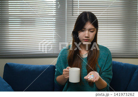 Young Asian Woman Hesitating to Take Medicine Pills While Holding a Mug at Home Young Asian Woman Hesitating to Take Medicine Pills While Holding a Mug at Home 141973262