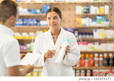 Female consultant pharmacist holds bottle of shampoo and offers to man client to purchase remedy Female consultant pharmacist holds bottle of shampoo and offers to man client to purchase remedy 141974177