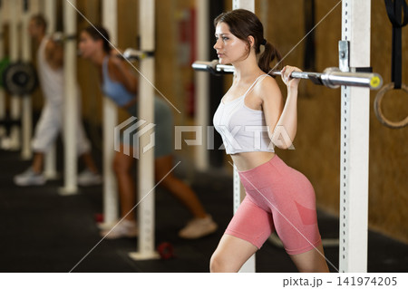 Focused girl in activewear exercising with barbell during group workout in gym 141974205