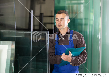 Experienced worker checks the quality of made window glass in an industrial workshop, marks the data in documents Experienced worker checks the quality of made window glass in an industrial workshop, marks the data in documents 141975583