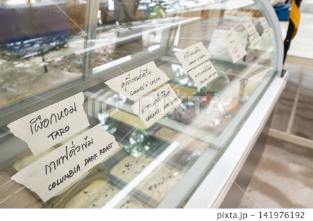 Glass ice cream freezer with handwritten Thai labels and English translations for assorted flavors including taro, coffee, vanilla, yogurt, passion fruit, and strawberry. Glass ice cream freezer with handwritten Thai labels and English translations for assorted flavors including taro, coffee, vanilla, yogurt, passion fruit, and strawberry. 141976192