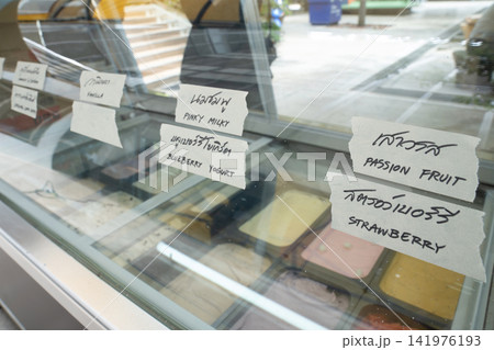 Glass ice cream freezer with handwritten Thai labels and English translations for assorted flavors including taro, coffee, vanilla, yogurt, passion fruit, and strawberry. Glass ice cream freezer with handwritten Thai labels and English translations for assorted flavors including taro, coffee, vanilla, yogurt, passion fruit, and strawberry. 141976193
