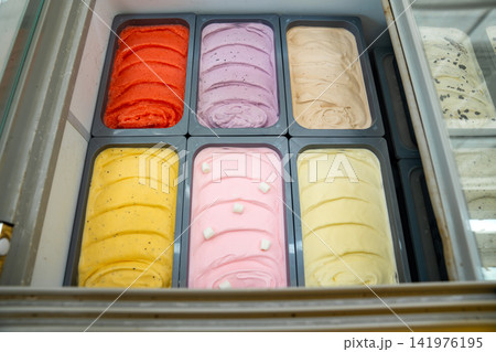 Various colorful ice cream flavors are neatly arranged in metal tubs inside a commercial freezer display. Rows of freshly churned gelato offer sweet dessert choices. Various colorful ice cream flavors are neatly arranged in metal tubs inside a commercial freezer display. Rows of freshly churned gelato offer sweet dessert choices. 141976195