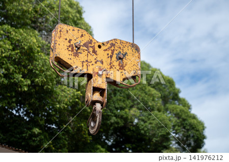 An old yellow crane sheave block with a steel hook hangs outdoors. An old yellow crane sheave block with a steel hook hangs outdoors. 141976212