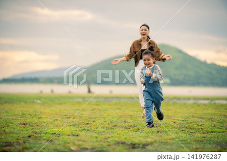 happy preschool child girl running with her mother on a grass field near river and mountain happy preschool child girl running with her mother on a grass field near river and mountain 141976287