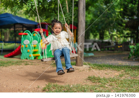 happy preschool child girl swinging on a wooden swing in a playground. happy preschool child girl swinging on a wooden swing in a playground. 141976300