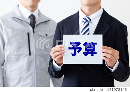 予算のホワイトボードを持つスーツを着た若い男性ビジネスマンと作業着のミドル男性 予算のホワイトボードを持つスーツを着た若い男性ビジネスマンと作業着のミドル男性 141978146