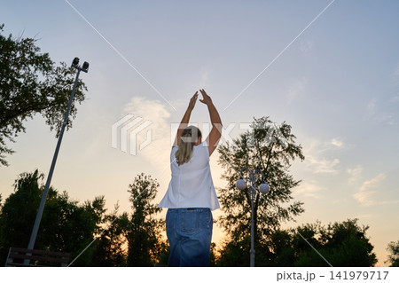 Woman Stretching Arms Overhead In Park At Sunset With Trees And Lamp Posts Against Evening Sky 141979717
