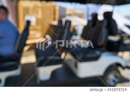 Empty Golf Cart Seats Parked Under Shade At Outdoor Venue With Blurred People And Wooden Backdrop Empty Golf Cart Seats Parked Under Shade At Outdoor Venue With Blurred People And Wooden Backdrop 141979724
