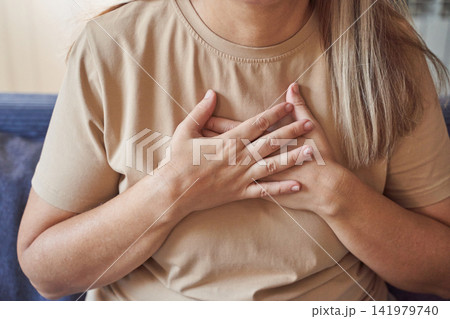 Woman Placing Hands Over Chest In Seated Pose On Sofa Indoors Wearing Beige Tee With Long Blond 141979740