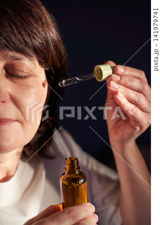 Mature Woman Applying Facial Serum With Dropper Near Closed Eyes Over Amber Bottle In Dim Studio 141979741
