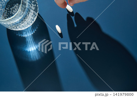 Glass Bottle Tablet And Hand Picking Pill Over Blue Surface Casting Long Hand Shadow With Sparkling Glass Bottle Tablet And Hand Picking Pill Over Blue Surface Casting Long Hand Shadow With Sparkling 141979800