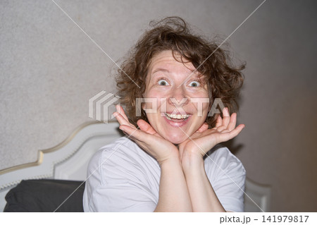 Woman Smiling With Wide Eyes And Both Hands Framing Face Beside Laptop In Bedroom, Expressing Woman Smiling With Wide Eyes And Both Hands Framing Face Beside Laptop In Bedroom, Expressing 141979817