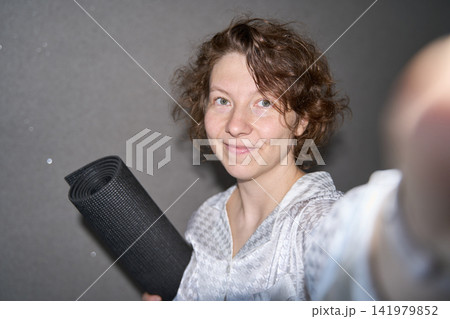 Smiling Woman Reaching Toward Camera While Holding Rolled Black Yoga Mat Indoors In Striped 141979852
