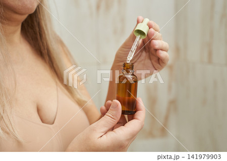 Woman Holding Amber Bottle With Green Cap And Pipette Preparing Serum For Application In Bathroom Woman Holding Amber Bottle With Green Cap And Pipette Preparing Serum For Application In Bathroom 141979903