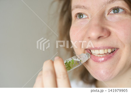Young Woman Smiling While Applying Lip Gloss From Glass Roller Bottle In Bright Neutral Indoor Young Woman Smiling While Applying Lip Gloss From Glass Roller Bottle In Bright Neutral Indoor 141979905