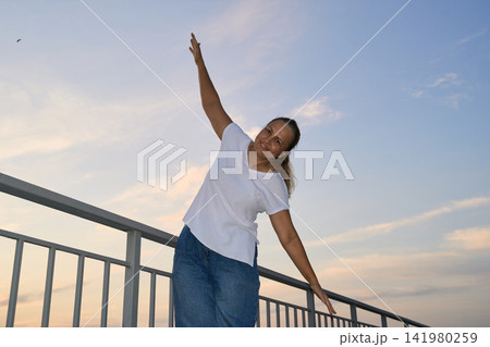 Adult Woman Posing With Raised Arm On Rooftop Railing At Sunset Under Pastel Sky 141980259