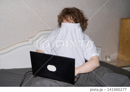 Woman Covering Face With White Shirt While Sitting Behind Open Laptop In Bedroom, Expressing Woman Covering Face With White Shirt While Sitting Behind Open Laptop In Bedroom, Expressing 141980277
