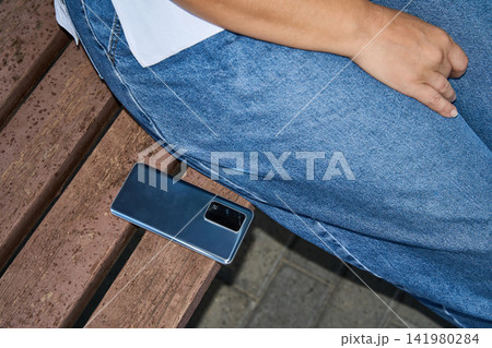 Smartphone Resting On Wooden Bench Beside Seated Woman In Denim Outfit At Park Walkway With Close Smartphone Resting On Wooden Bench Beside Seated Woman In Denim Outfit At Park Walkway With Close 141980284