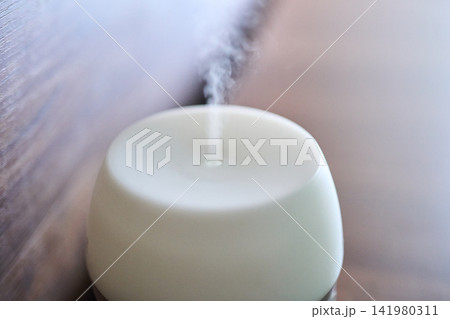 Aroma Diffuser Releasing Thin White Mist On Wooden Tabletop Closeup In Indoor Living Space Aroma Diffuser Releasing Thin White Mist On Wooden Tabletop Closeup In Indoor Living Space 141980311
