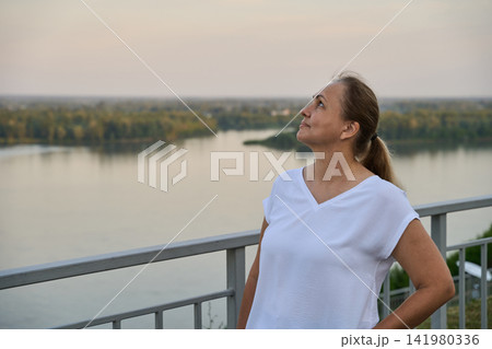 Adult Woman Standing Beside Railing And Looking Upward Over River Landscape In Soft Evening Light Adult Woman Standing Beside Railing And Looking Upward Over River Landscape In Soft Evening Light 141980336