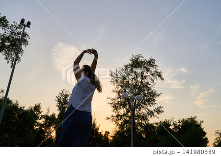 Woman Stretching Arms Overhead In Side Silhouette At Sunset Among Trees And Park Lights 141980339
