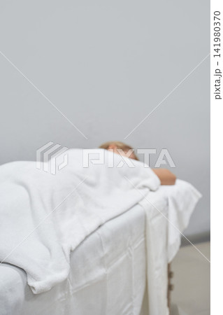 Person Resting Face Down Under White Blanket On Massage Table In Minimalist Treatment Room 141980370