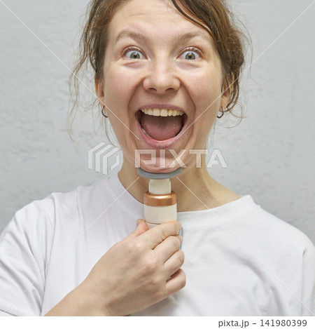 Woman Holding Electric Shaver At Throat Expressing Wide Open Mouth Excitement Against Plain Light Woman Holding Electric Shaver At Throat Expressing Wide Open Mouth Excitement Against Plain Light 141980399