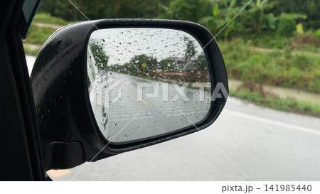 Close up view of a car side mirror covered in raindrops, reflecting a wet road during rainy weather. Road with a stream running alongside it in a rural area. Close up view of a car side mirror covered in raindrops, reflecting a wet road during rainy weather. Road with a stream running alongside it in a rural area. 141985440