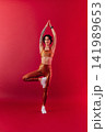 Full length of a young, slim woman in golden sportswear raising her hand up while standing on one leg 141989653