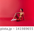 Young smiling woman in golden fitness attire sitting on a crimson background and looking up 141989655