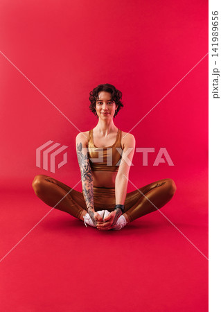 Young slim woman in golden fitness attire sitting on a crimson background Young slim woman in golden fitness attire sitting on a crimson background 141989656