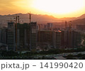 Tall residential buildings under construction with multiple cranes rising above the city skyline during a golden sunset near mountains in China 141990420