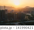 Tall residential buildings under construction with multiple cranes rising above the city skyline during a golden sunset near mountains in China 141990421